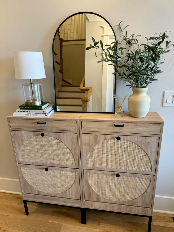 Cane entry cabinet styled with a black arch mirror, table lamp, stacked books, and a cream vase with olive branches.