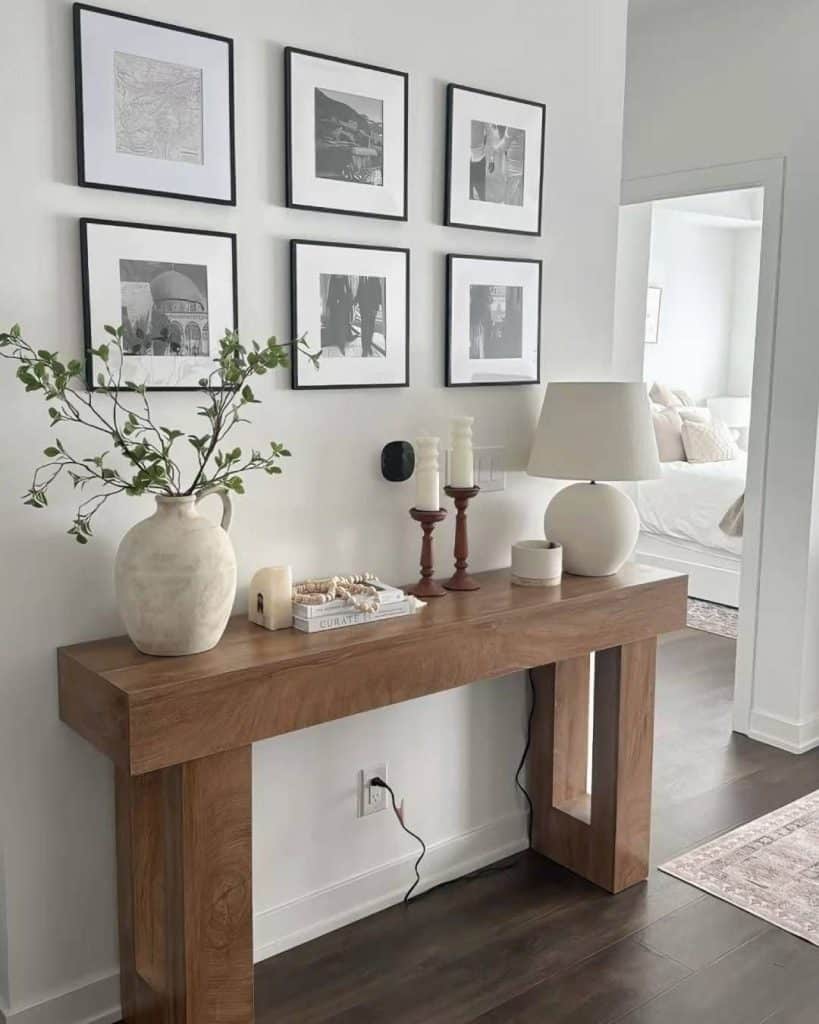Chunky natural wood console table below a 2x3 gallery wall of black-framed black and white prints, styled with a cream jug vase, wooden candlesticks, beaded garland, and a white globe lamp.