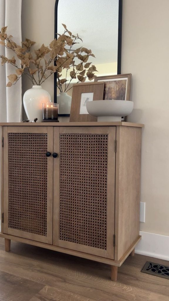 Light cane cabinet topped with a tall black arch mirror, white ceramic vase with dried fall branches, glass candle, small framed prints, and a white bowl.
