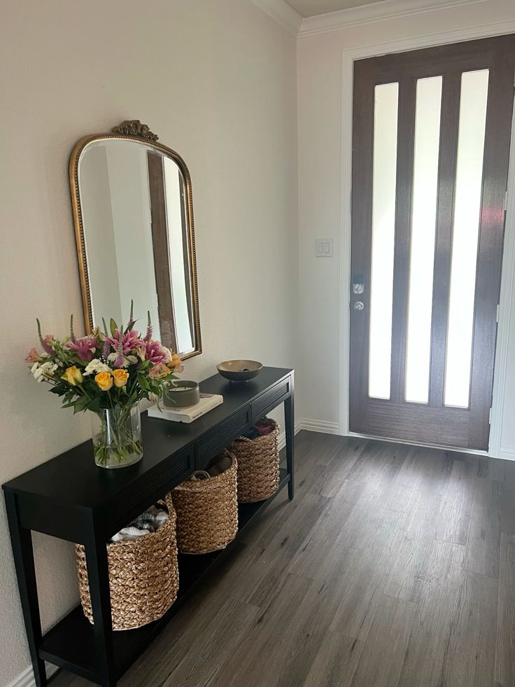 Black entry console with ornate tall gold arch mirror, glass vase with colorful fresh flowers, small ceramic bowl, and two woven baskets on the lower shelf.