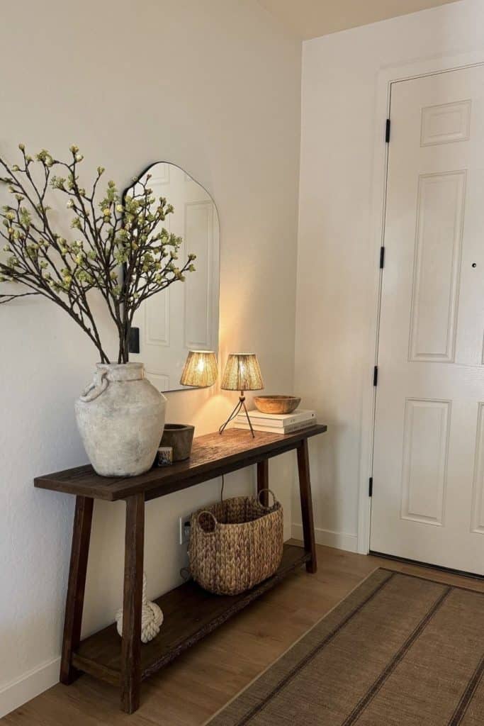 Dark wood rustic bench entry table with arch black mirror, large terracotta vase with spring branches, two tripod rattan lamps, stacked books, wooden bowl, and a woven basket below.