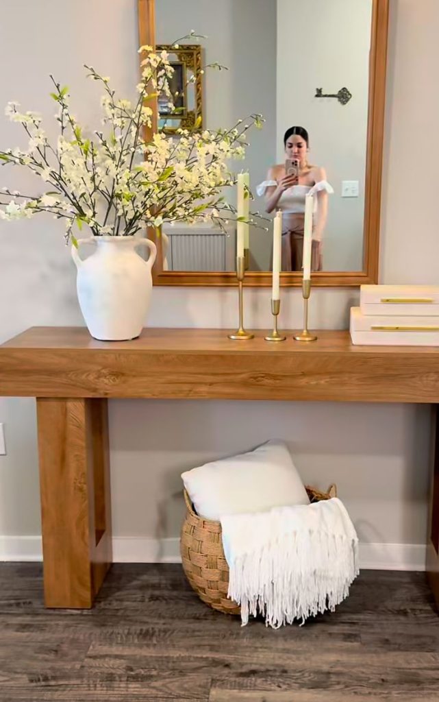 Natural wood entry console with gold rectangular mirror reflection, white pitcher vase with white blossom branches, three gold taper candlesticks, stacked books, and a woven basket with a white throw below.