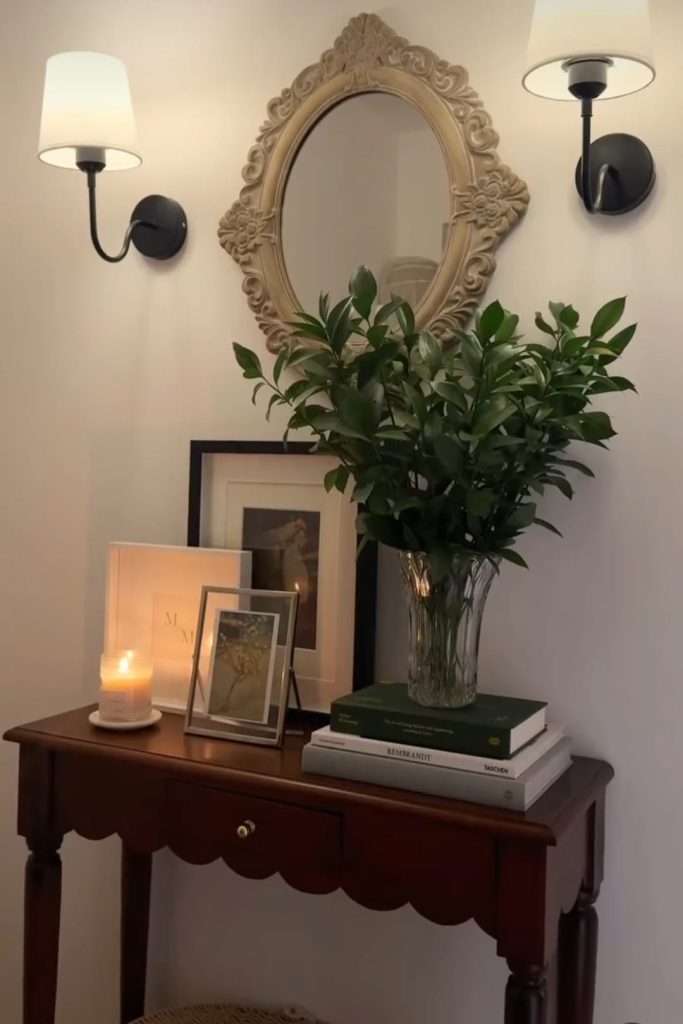 Vintage dark wood scalloped entry table with ornate cream oval carved mirror, two black wall sconces, stacked books, multiple framed art prints, crystal vase with lush green stems, and a pillar candle.