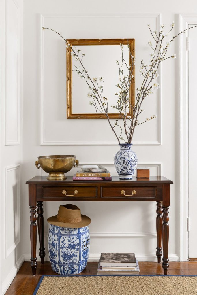 Dark mahogany entry table with gold square framed mirror, blue and white chinoiserie vase with white blossom branches, brass bowl, stacked books, and a blue and white ceramic garden stool below.