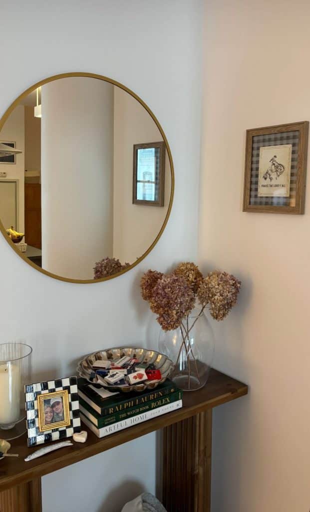 Dark walnut fluted console table with round black framed mirror, tall woven vase with dried eucalyptus, stacked books, small shell dish, candle diffuser, and a boucle round stool below.