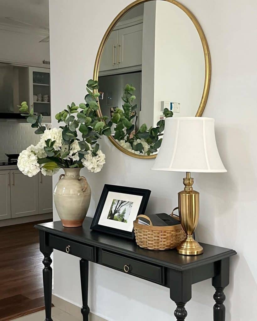 Black entry table with a large round gold mirror, white hydrangeas in a cream vase, gold lamp, framed photo, and woven basket.