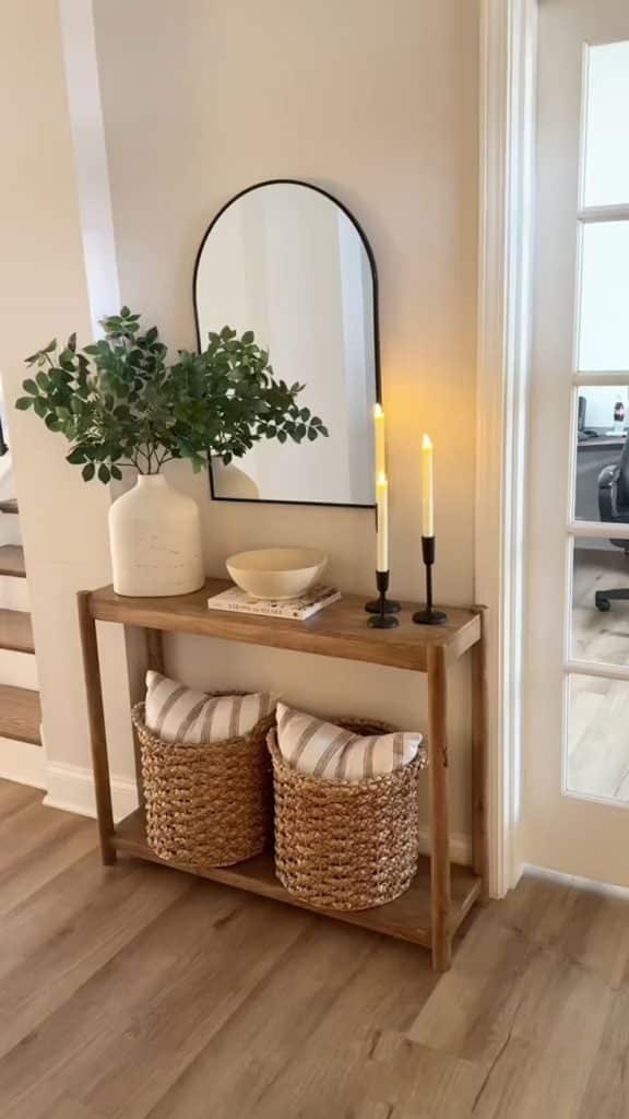 Natural wood entry table with black arch mirror, white vase with greenery, bowl, candles on black holders, and two woven baskets underneath.