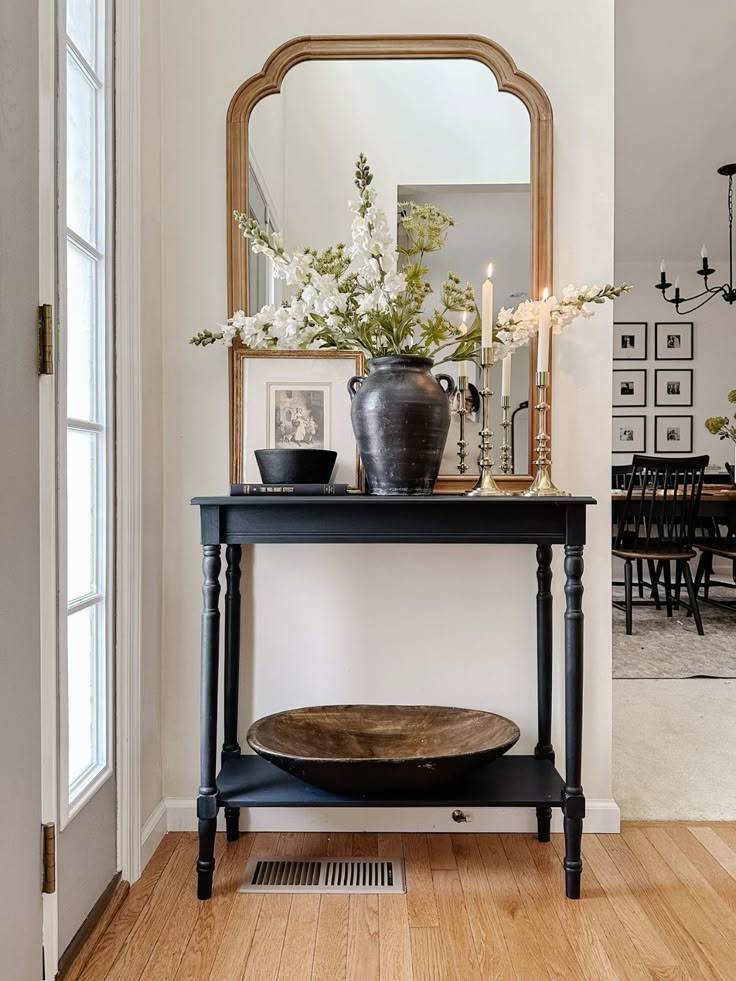 Black entry console with large carved wood arch mirror, dark urn with white flowers, brass candlesticks, framed art, and a wood dough bowl on the lower shelf.