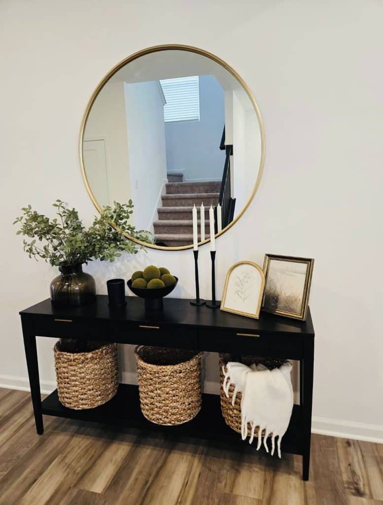 Black entry table with large round gold mirror, greenery in dark vase, taper candles on black holders, framed art prints, and three woven baskets on lower shelf.