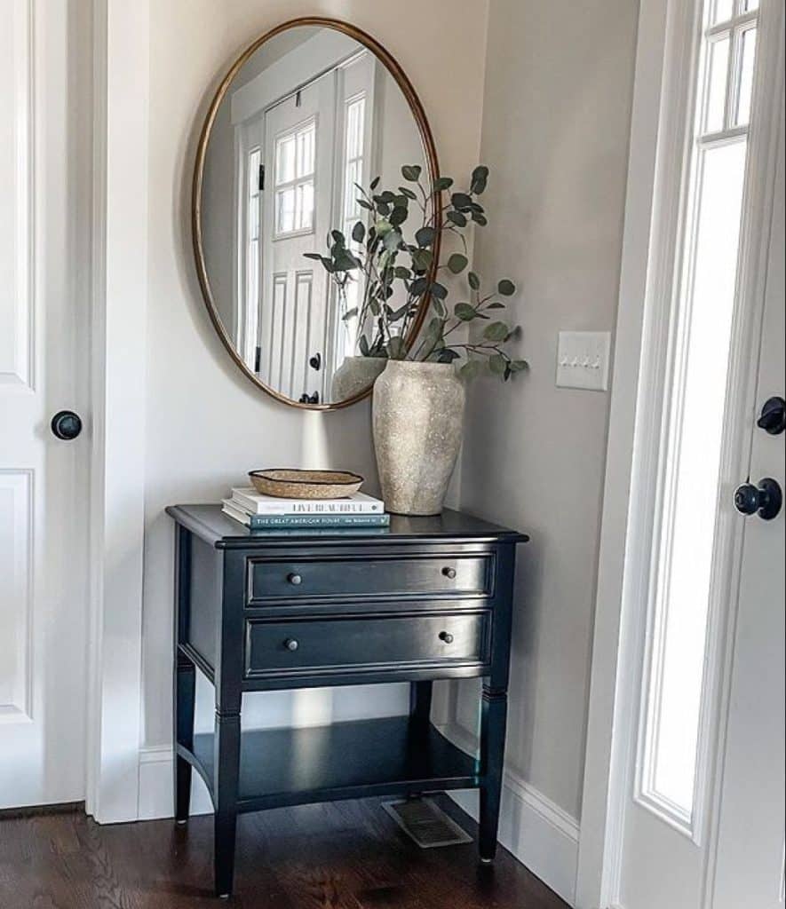 Small dark entryway cabinet between two white doors, styled with a round gold mirror, stacked books, woven tray, and tall cream vase with eucalyptus.