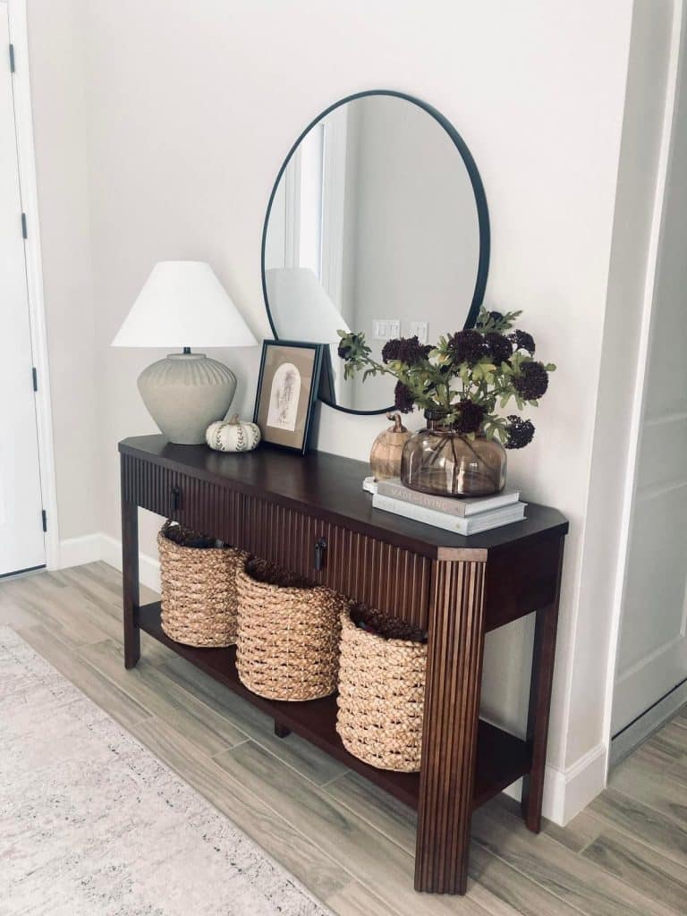 Dark wood fluted console table with round black oval mirror, ceramic lamp, small pumpkin, framed art, glass vase with dark flowers, and three woven baskets below.