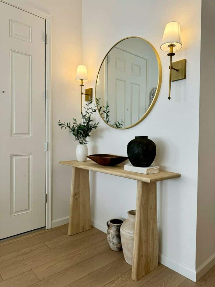 Light wood entry table near front door with round gold mirror, two gold wall sconces, white vase with greenery, dark ceramic pot, and rustic jugs below.
