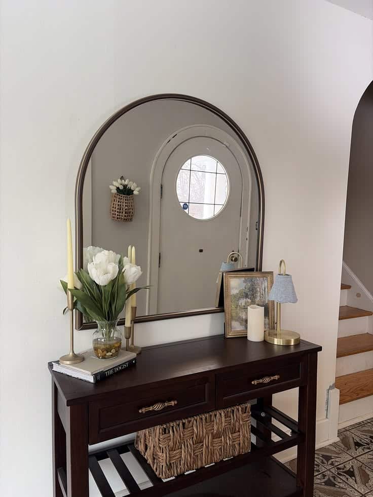 Dark wood entry table with large arch bronze mirror, white tulips in glass vase, gold candlesticks, framed art, small lamp, and woven basket below.