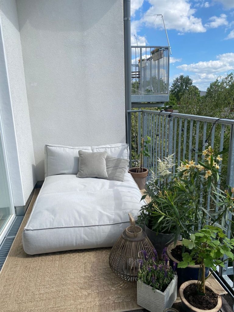 Minimalist white balcony with oversized floor cushion lounger, rattan lantern and potted flowering plants