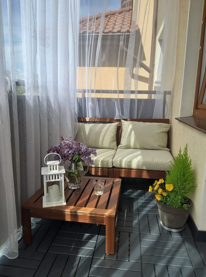 Small balcony with timber corner sofa set, cream cushions, lantern with candle, fresh lilac flowers and dark tile deck