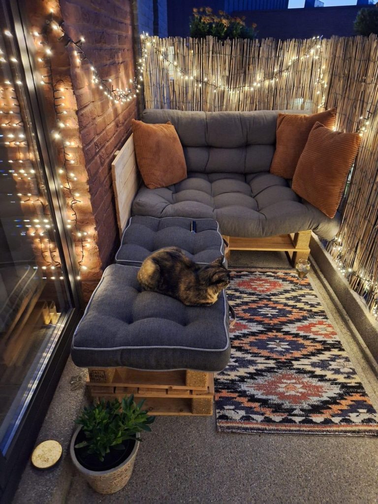 Cozy small balcony at night with pallet sofa, orange cushions, fairy lights and bamboo screen privacy