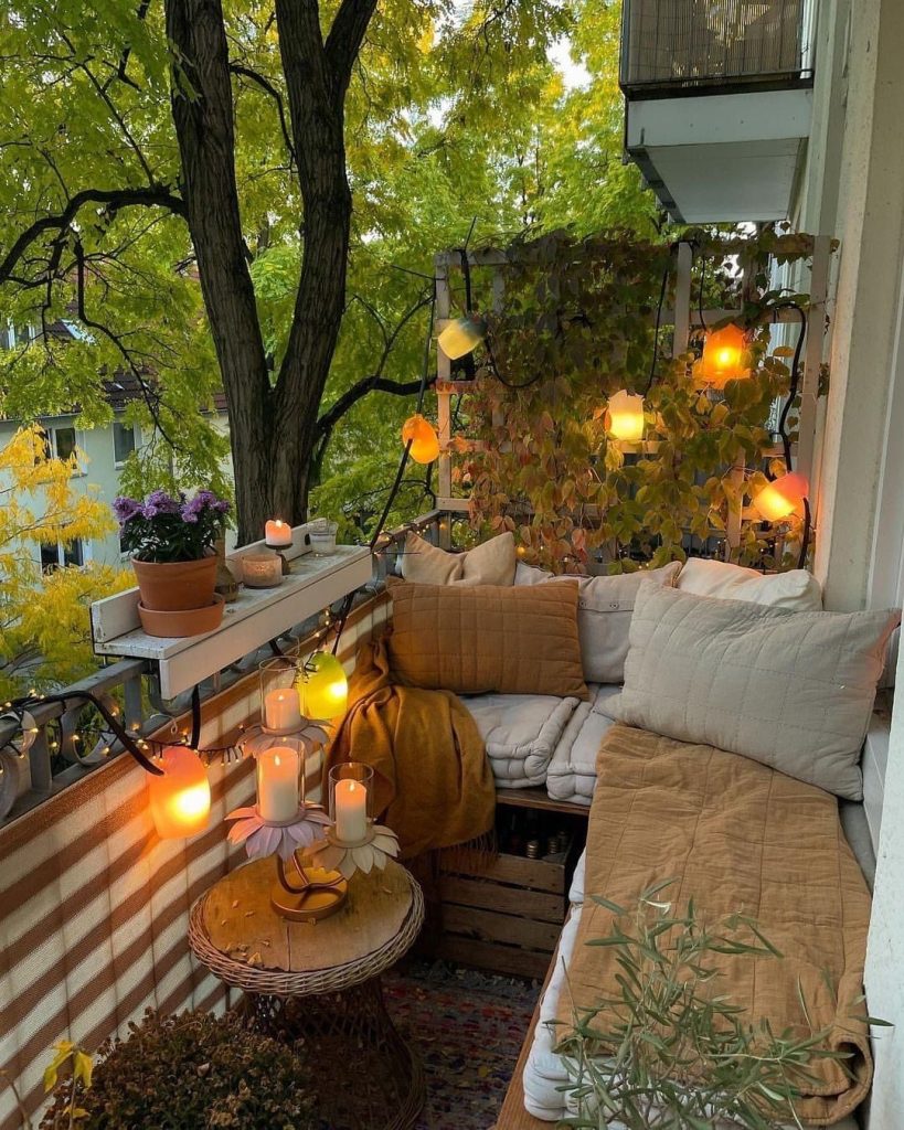 Cozy balcony corner sofa with warm candles, globe string lights, autumn foliage and a wicker side table