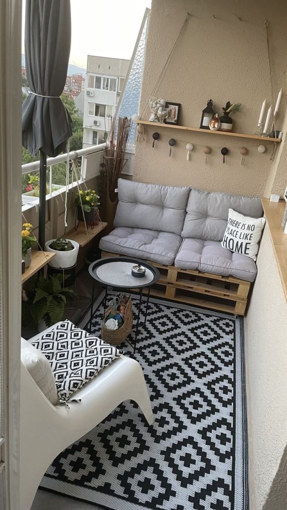 Small apartment balcony with pallet sofa, grey cushions, black and white rug and wall shelf with decor