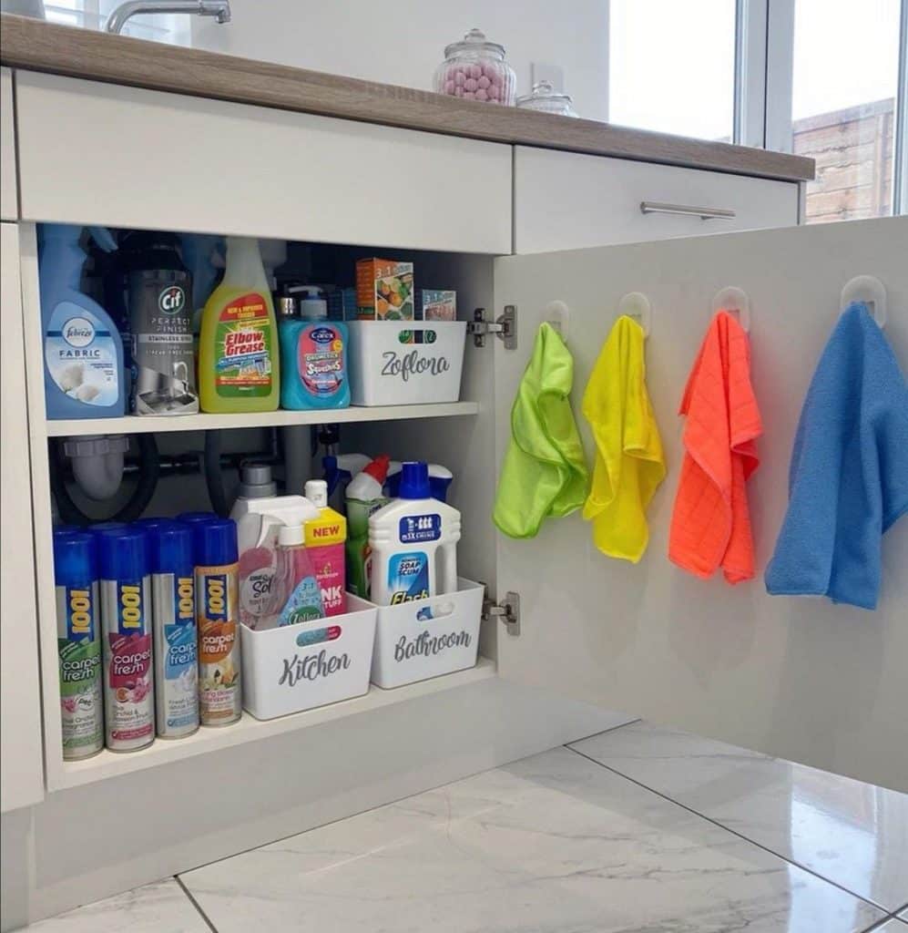 Under kitchen sink with two shelves of cleaning products in labeled white bins and four colorful microfibre cloths hanging on hooks on the inside of the cabinet door