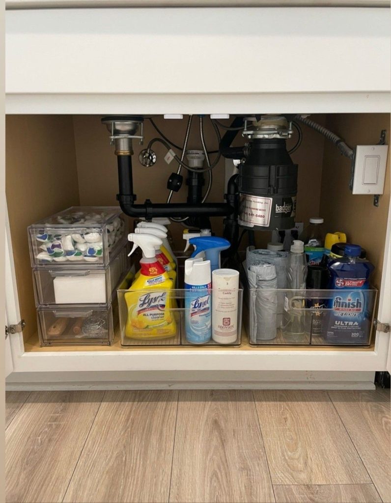 Under kitchen sink cabinet with a garbage disposal unit showing clear acrylic pull out mini drawers on the left holding dish tablets and sponges, and larger clear bins on the right grouping cleaning sprays, dish pods and laundry products