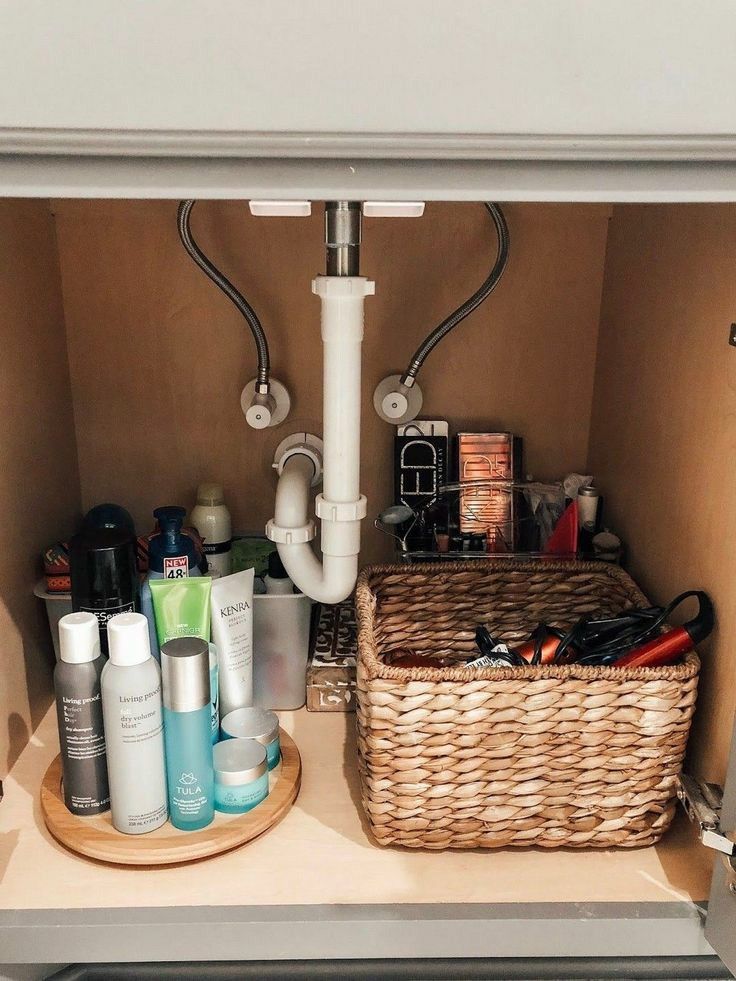 Under bathroom sink with a wooden lazy susan holding hair care and skincare products on the left side of the pipes and a large wicker basket holding hair tools on the right side