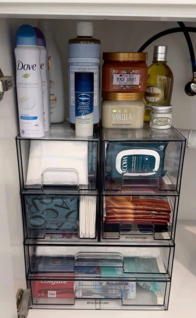 Under bathroom sink with a column of six stacked acrylic pull out drawers holding cotton rounds, cotton buds, menstrual products, face wipes and toothbrushes, with body care products displayed on top