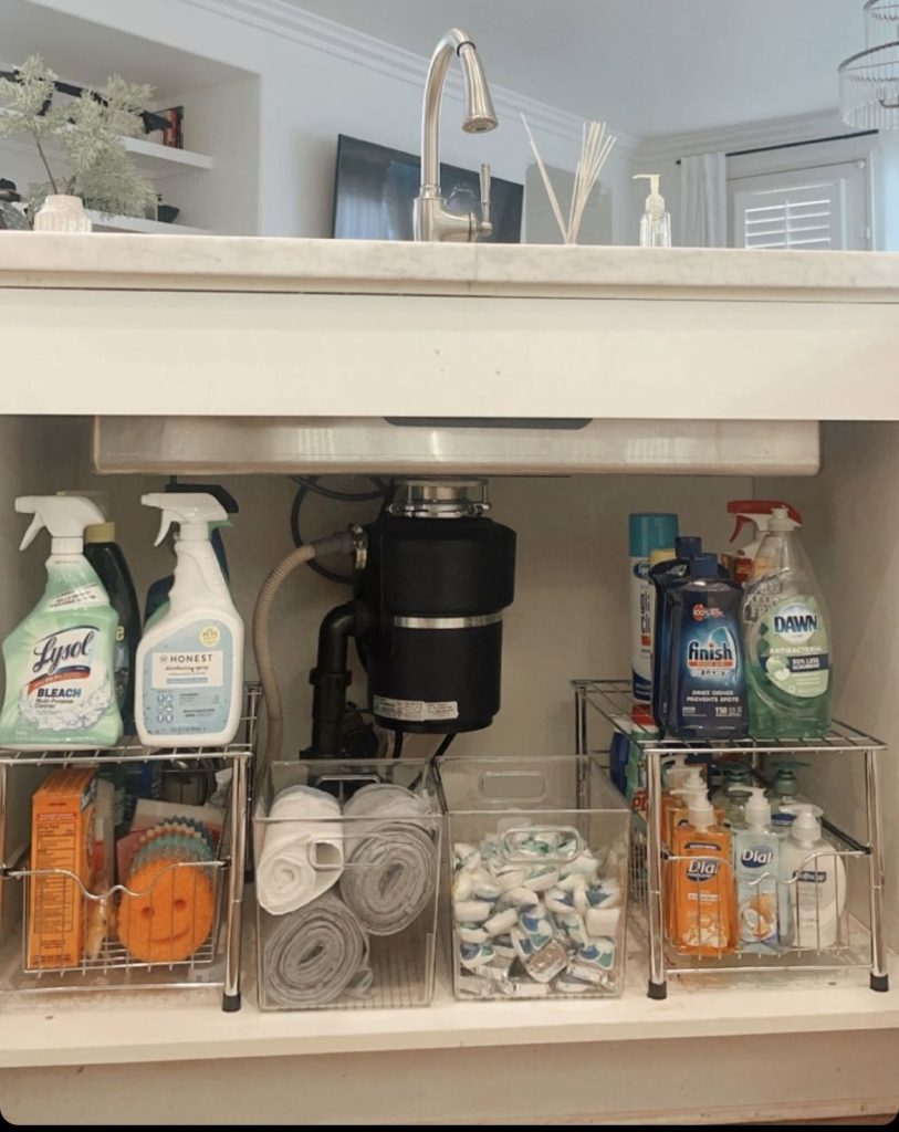 Under kitchen sink with a garbage disposal showing two wire shelf units on each side holding cleaning sprays on top and clear and wire bins below holding sponges, dish soap, laundry products and dishwasher pods