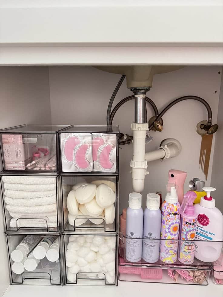 Under bathroom sink with a stack of six dark framed acrylic pull out drawers on the left holding eye patches, washcloths, bath soaps and cotton rounds, and a clear shelf unit on the right holding hair products and skincare