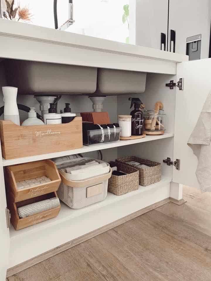Under kitchen sink cabinet with two built in shelves holding labeled bamboo boxes, fabric and wicker baskets and glass cleaning spray bottles in a warm natural style