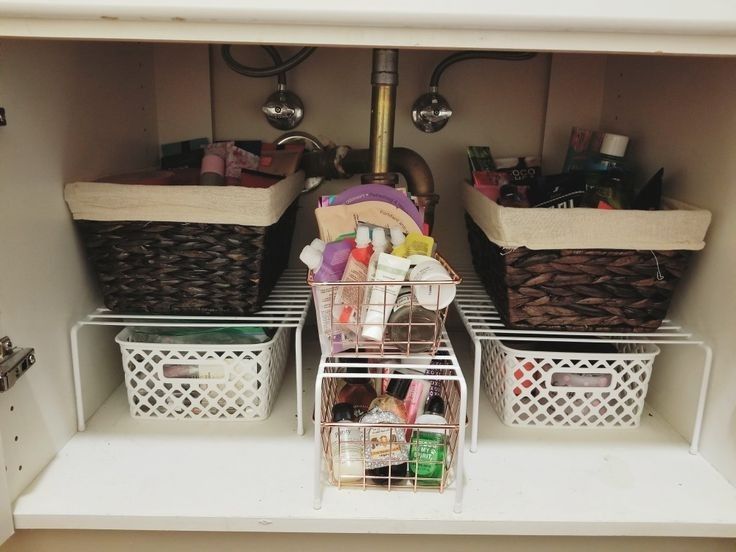 Under bathroom sink with two large wicker storage baskets with linen liners on raised white wire shelf risers, and a smaller wire basket in the center holding beauty products