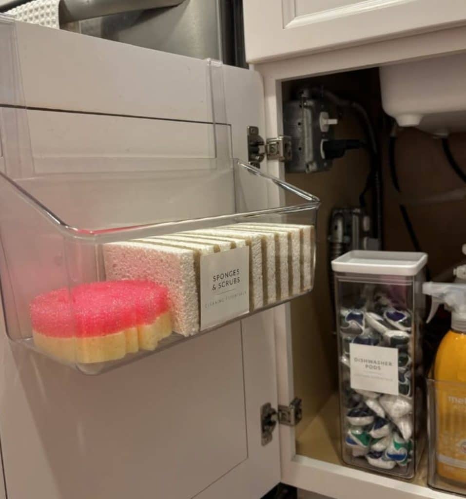 Clear acrylic over the door bin mounted on the inside of a kitchen cabinet door holding labeled sponges and scrubs, with a tall clear container of dishwasher pods and cleaning sprays visible inside the cabinet