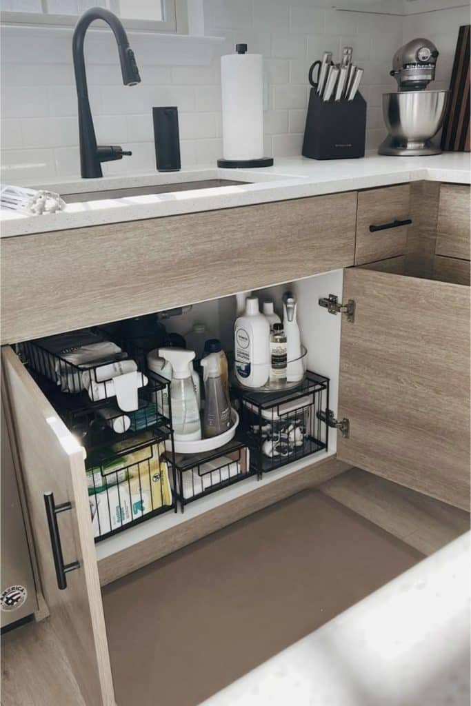 Under kitchen sink cabinet with black wire pull out baskets on both sides holding dish soap, cleaning sprays, sponges and a lazy susan turntable in the center
