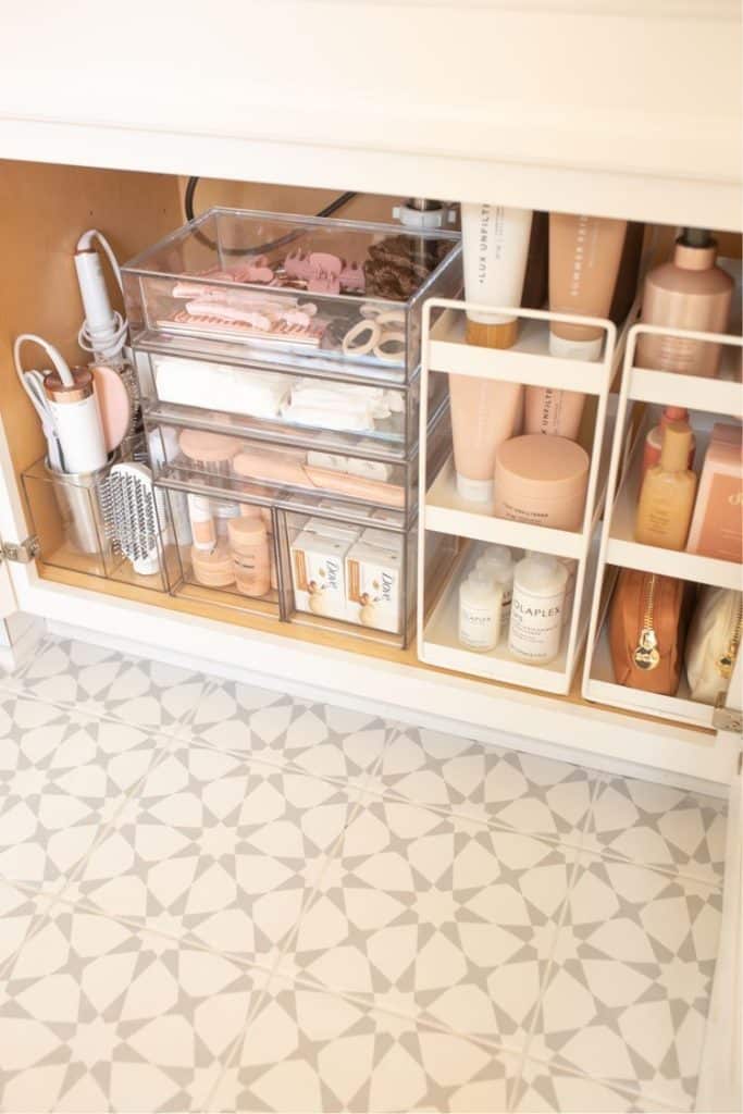 Under bathroom sink with stacked clear acrylic drawers on the left holding hair clips and cosmetics, and a white two tier shelf unit on the right holding hair products and skincare