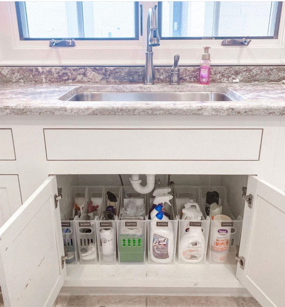 Under kitchen sink cabinet with a row of tall labeled plastic organizer bins in a row holding sponges, gloves, specialty products, a swiffer, bathroom cleaner, all purpose spray and disinfecting wipes