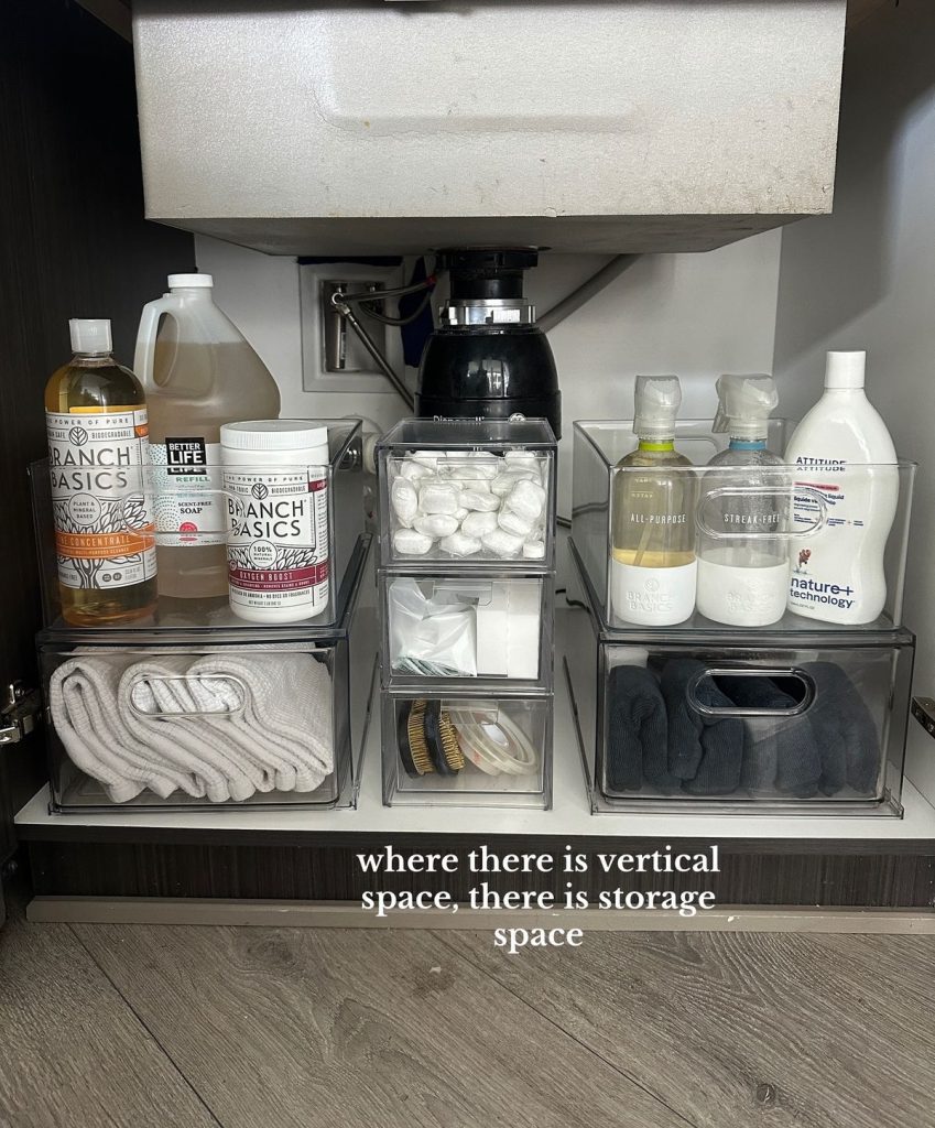 Under kitchen sink showing stacked clear acrylic bins on both sides of a garbage disposal holding cleaning concentrate bottles, dish tablets, dish cloths and cleaning gloves