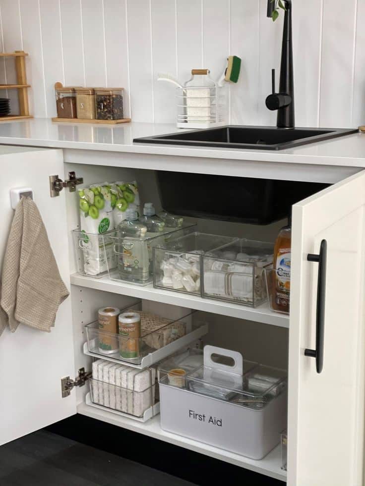 Under kitchen sink with a white built in shelf creating two storage levels, clear bins holding dish pods and sponges on the top shelf and a white first aid box and paper towels on the lower shelf