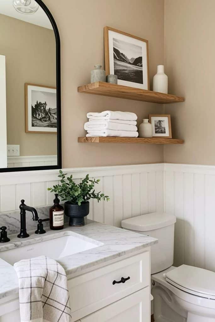 Neutral guest bathroom with wooden floating shelves styled with folded white towels, ceramic vases, and black and white framed art above a white vanity with marble countertop and black fixtures.