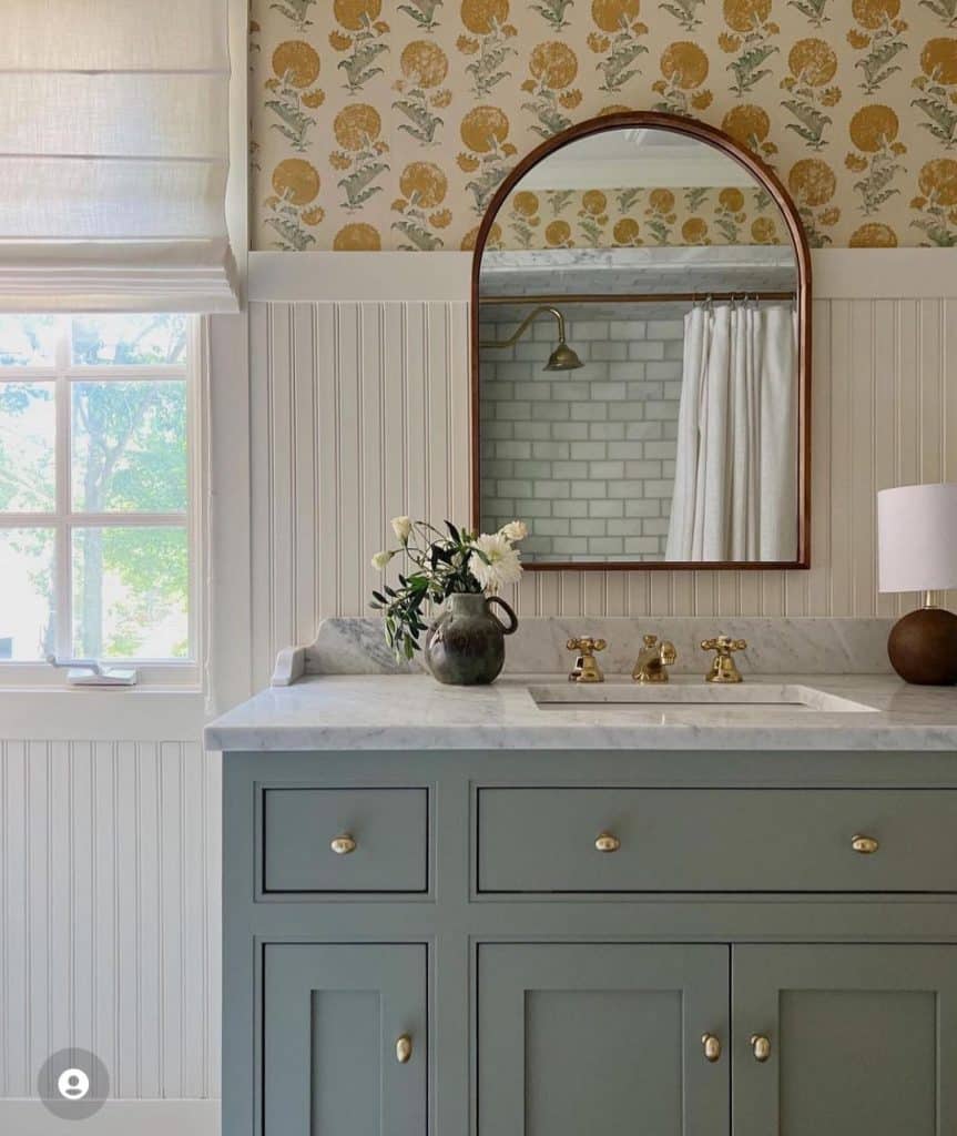 Charming guest bathroom with bold yellow and green floral wallpaper above white beadboard, a dusty teal vanity with gold oval hardware, a marble countertop, gold cross-handle faucets, an arched walnut-framed mirror, a dark ceramic vase with white flowers, and a small table lamp.