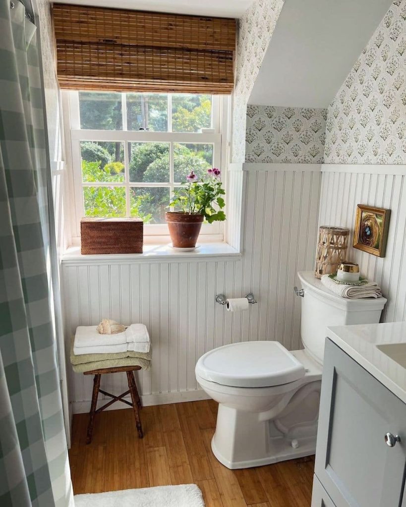 Cottage-style small guest bathroom with white beadboard wainscoting, delicate green and white floral wallpaper, a bamboo roman blind on the window, a potted pink geranium on the windowsill, a wicker rattan box, a small rustic wooden stool with stacked towels, and a gingham shower curtain.