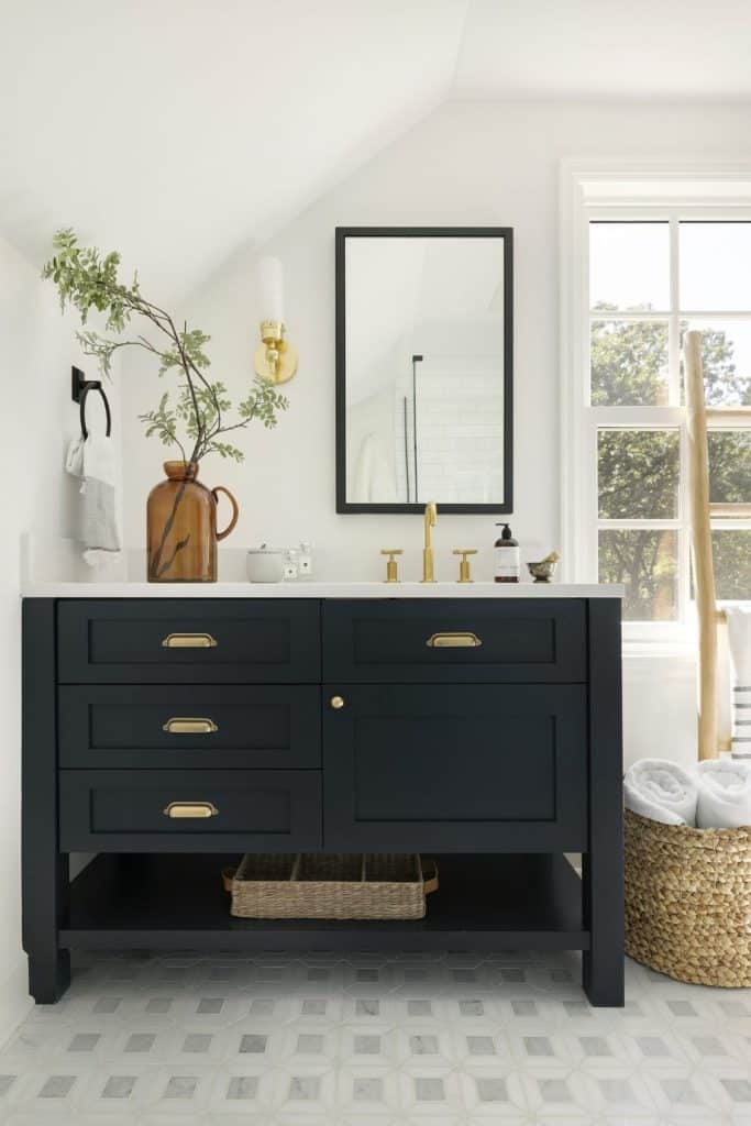 Stylish guest bathroom with a freestanding dark navy vanity with brass cup pull hardware, a white quartz countertop, a black-framed rectangular mirror, a warm gold wall sconce, a tall amber glass vase with greenery, and a large woven basket holding white towels beside a window.