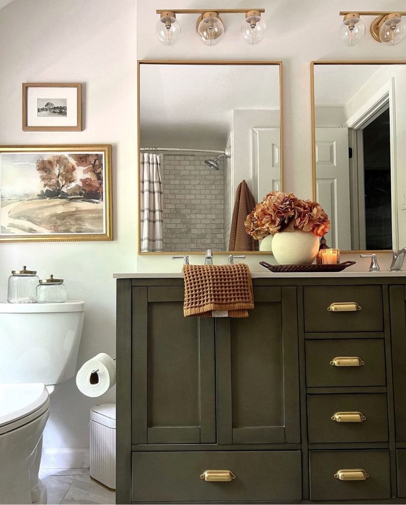 Warm guest bathroom with an olive green painted double vanity, brass cup pull hardware, two gold-framed rectangular mirrors, globe globe bathroom lighting in brass, a large vintage landscape painting on the wall, a white ceramic vase with dried orange hydrangeas, and a rattan tray with a candle on the countertop.