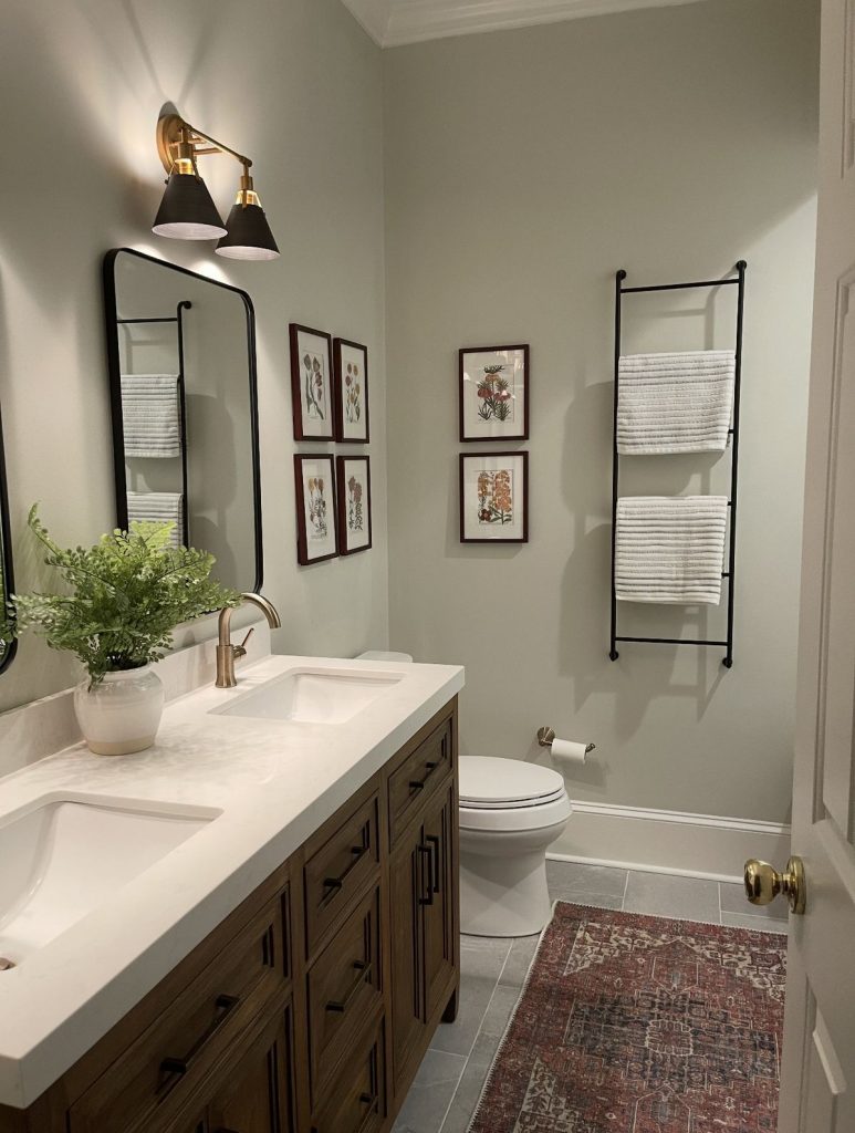 Refined sage green guest bathroom with a timber-stained vanity, black-framed curved mirror, dual-arm black and brass wall sconce, a wall-mounted black metal towel ladder with white stripe towels, four framed vintage botanical prints arranged in a grid, a white ceramic pot with a fern plant, and a vintage Persian rug on grey tile floors.