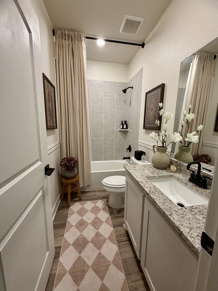 Narrow guest bathroom with cream wainscoting, a long linen shower curtain hung high from a black rod, a granite-look countertop on a white vanity, matte black hardware and fixtures, a harlequin patterned runner rug, framed vintage art prints on the walls, and a ceramic pot with dried hydrangeas and magnolia stems on the vanity.