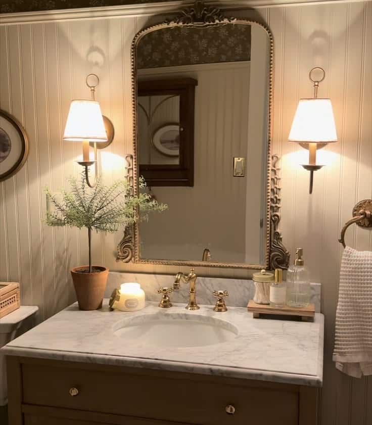 Romantic vintage guest bathroom with warm beige beadboard walls, an ornate antique gold-framed mirror, dual candle-style wall sconces with white lampshades, a marble countertop with a round undermount sink, polished brass cross-handle faucets, a terracotta pot with a small herb plant, a lit candle, a glass soap dispenser, and a waffle-weave hand towel.