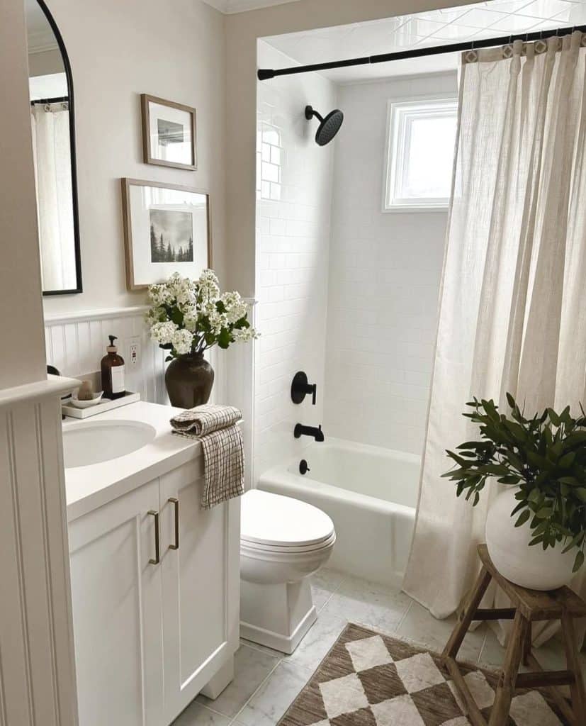 Fresh all-white guest bathroom with white subway tile walls, a white vanity with brass hardware, matte black shower fixtures and curtain rod, a linen shower curtain, a black arched mirror, beadboard wainscoting, framed forest photography prints, a dark ceramic vase with white hydrangeas, a checkered woven bath mat, and a large potted plant on a wooden step stool.