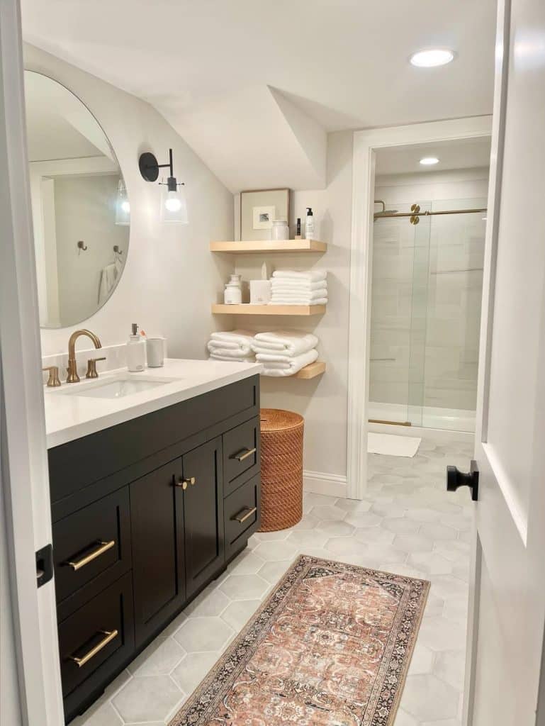 Modern guest bathroom with a dark charcoal vanity, brass hardware, round mirror, gold sconce lighting, hexagonal floor tiles, a Persian runner rug, and light wood floating shelves stacked with white towels.