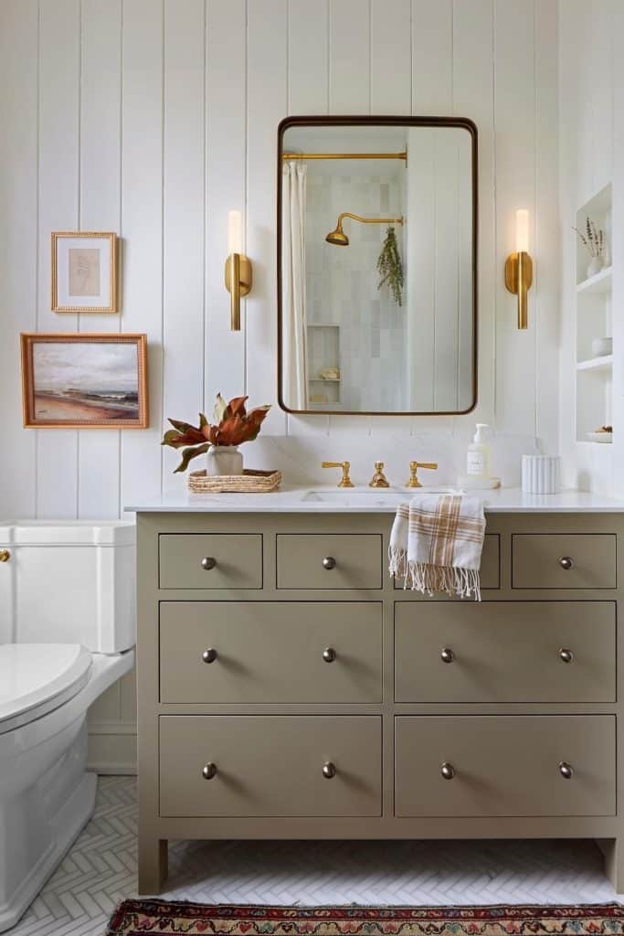 Elegant guest bathroom with a sage green painted vanity, brass faucet and sconce lighting, an arched gold-framed mirror, shiplap paneling walls, a plaid hand towel, and small framed landscape art.