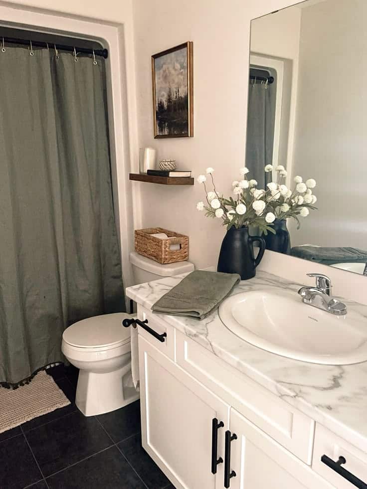 Budget-friendly white guest bathroom with dark tile floors, a sage green shower curtain, marble-look countertop, black hardware accents, a small floating shelf with decor, a wicker basket on the toilet tank, and a dark ceramic vase with white flowers.