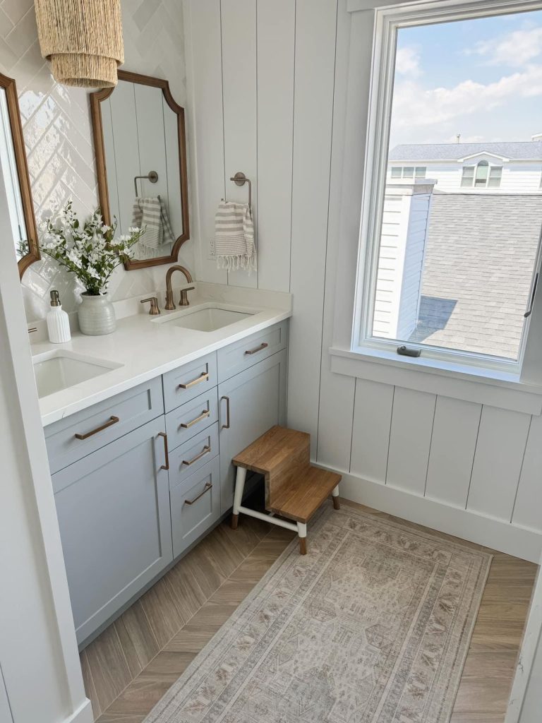 Bright coastal-style guest bathroom with a powder blue double vanity, bronze hardware, a wood-framed arched mirror, shiplap paneling, herringbone wood-look floor tiles, striped hanging hand towels, and white flowers on the countertop.