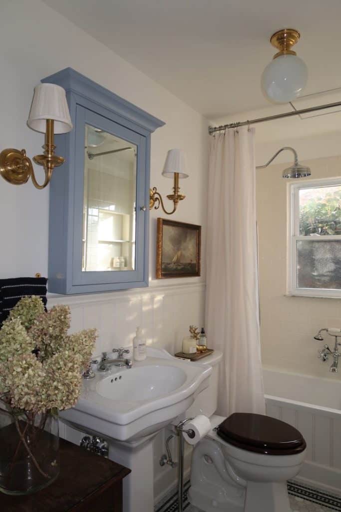 Vintage-style guest bathroom with a powder blue medicine cabinet, brass candle sconces, a pedestal sink, clawfoot bath with curtain, beadboard wainscoting, globe ceiling light, dark timber toilet seat, and dried hydrangeas in a glass vase.